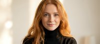 Young redhead woman with long wavy hair wearing black turtleneck sweater smiling at camera in modern indoor setting for lifestyle and portrait photography.