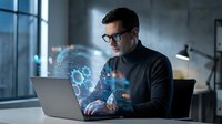 Professional businessman with glasses working on laptop with holographic data visualization and digital interface projections in modern office environment.