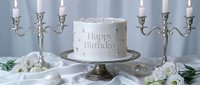 Elegant white birthday cake with silver star decorations and Happy Birthday inscription, flanked by silver candelabras and white flowers on silk.