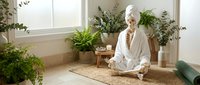 Mature woman with white hair in white linen clothing meditating among potted plants and greenery by bright window, wellness and mindfulness practice.