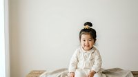 Young Asian child, approximately 1-2 years old, wearing a light beige long-sleeve shirt, sitting on a bed with neutral bedding against a white wall background.