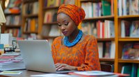 Young African woman in bright orange headwrap and traditional dress working on laptop in library setting, wearing blue beaded necklace. Professional student studying.
