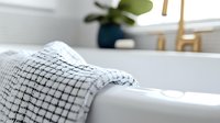 Soft white waffle weave cotton towel draped over bathtub edge in modern minimalist bathroom with green plant decor, creating serene spa-like atmosphere.