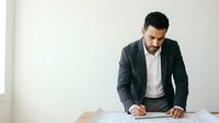 Young Middle Eastern male professional in dark blazer and white shirt reviewing documents at desk against white background.