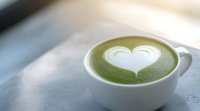 Creamy heart-shaped latte art on frothy matcha green tea served in white ceramic cup against blurred bright background. For cafe and beverage menus.