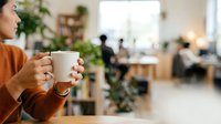Closeup of woman's hands holding warm mug in modern office workspace with blurred colleagues working at desks behind, ideal for corporate wellness and workplace culture content.