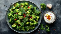 Fresh steamed broccoli florets seasoned with chili flakes and garlic, served on dark plate with creamy yogurt dip, overhead view on concrete background.