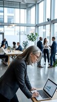 Middle-aged woman with long gray hair and glasses wearing dark business suit working on laptop in modern office with colleagues collaborating in background by windows.