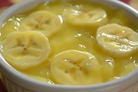 Fresh banana slices floating in smooth yellow pudding dessert, close-up view showing creamy texture and ripe fruit in white ceramic bowl.
