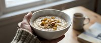Person in cozy sweater holding bowl of oatmeal porridge with nuts and dried fruit by window, healthy breakfast scene for wellness and nutrition content.