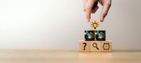 Hand placing lightbulb icon on wooden blocks with business symbols representing innovation and creative problem-solving in corporate strategy.