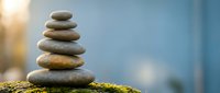 Balanced stack of smooth river stones on moss-covered surface with soft sunlight and blue sky background creating peaceful zen meditation atmosphere.