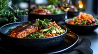Grilled salmon fillet with fresh green salad and vegetables served in dark ceramic bowl on black plate, moody restaurant atmosphere with bokeh lights.