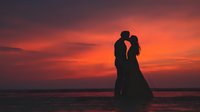 Romantic couple silhouette embracing at sunset beach with dramatic orange and pink sky reflecting on calm ocean water creating intimate wedding moment.