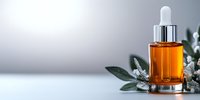 Amber glass dropper bottle with essential oil or serum against soft gray background, decorated with white flowers and green leaves. Luxury skincare concept.