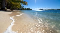 Tranquil tropical beach with gentle waves washing onto golden sand, palm trees swaying in breeze, crystal clear turquoise water and distant islands under blue sky.