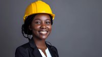 Confident African American female construction professional wearing yellow hard hat and business suit smiles warmly against dark background.