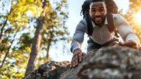 Joyful African American man with dreadlocks climbing rock in forest during golden hour sunset, wearing casual business attire.