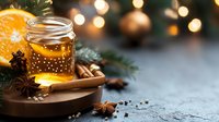 Glass jar of honey with orange slice, cinnamon sticks, star anise and winter spices against blurred Christmas lights background on dark stone surface. Cozy winter mood.