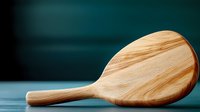 Minimalist wooden spoon lying on dark surface with blurred teal background. Natural kitchen utensil with smooth texture and curved design for cooking and serving.