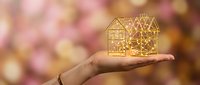 Hand holding golden wireframe house model against warm bokeh background for real estate investment and property development concepts.
