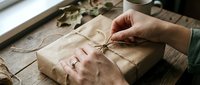 Hands tying twine around kraft paper wrapped gift box on wooden surface with dried foliage, suitable for holiday packaging and gift wrapping design projects.