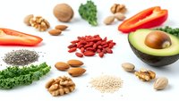 Healthy superfood ingredients including avocado, nuts, seeds, goji berries and fresh vegetables arranged on white background for nutritious meal planning.