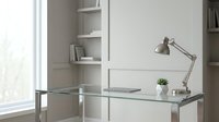Modern minimalist home office workspace with glass desk, adjustable metal lamp and white shelving unit creating clean professional environment.
