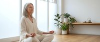 Peaceful mature woman with gray hair meditating in lotus position on wooden floor in bright modern room with plants for wellness and mindfulness practice.