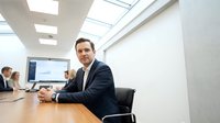 Professional businessman in dark suit sitting at modern conference table during corporate meeting with colleagues in bright office boardroom.