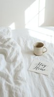 Cozy morning bed scene with ceramic mug and stay home calligraphy card on white linen, soft natural sunlight, minimal hygge lifestyle concept.