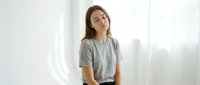 Young Caucasian woman in casual gray t-shirt sitting peacefully with eyes closed in bright minimalist interior with natural sunlight streaming through window.