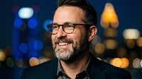 Middle-aged Caucasian businessman with glasses and beard smiling confidently against blurred city lights background at night for corporate portraits.
