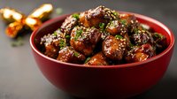 Glazed Asian meatballs garnished with sesame seeds and green onions in red ceramic bowl, dark background with warm lighting creates appetizing mood.