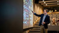 Professional businessman presenting data analytics on large screen in modern conference room with colleagues watching from tiered seating area.