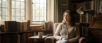 Middle-aged woman relaxing in cozy library reading nook with books, warm natural lighting from large windows, comfortable armchair for literary lifestyle.