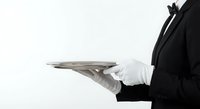 Professional waiter in black uniform holding silver serving tray with white gloves against clean white background for hospitality service concepts.
