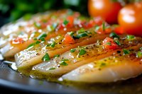 Fresh raw fish fillets seasoned with herbs, olive oil, cherry tomatoes and chives on black plate, close up view with selective focus for culinary use.