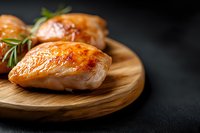 Grilled chicken breast with golden-brown crust and rosemary garnish on round wooden cutting board against dark background, close up view of juicy poultry.