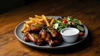 Grilled chicken wings with golden french fries and fresh salad served on dark plate. Restaurant meal with crispy potatoes and mixed greens ready for dining.