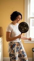 Happy young Hispanic woman cooking in sunny kitchen holding cast iron skillet wearing casual clothes and patterned pants for home lifestyle content.