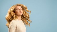 Young blonde woman with flowing wavy hair in motion wearing beige knit sweater against blue background. Natural beauty lifestyle portrait for wellness concepts.