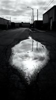 Monochrome industrial alley with puddle reflection of cloudy sky, abandoned warehouse district, urban decay and gritty street photography for editorial and artistic projects.
