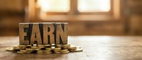Wooden block letters spelling earn stacked on pile of gold coins on wooden surface with blurred window background, representing income growth and financial success.