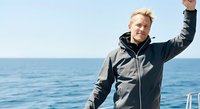 Caucasian male sailor in dark waterproof jacket raises fist triumphantly on boat deck with calm ocean and clear sky behind him, celebrating success and achievement at sea.
