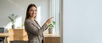 Young professional businesswoman in gray suit pointing and presenting in modern office space with natural lighting and plants.
