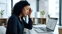 Stressed African American businesswoman with headache working on laptop in modern office environment experiencing workplace fatigue and mental exhaustion.
