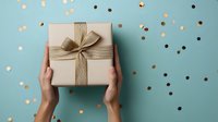 Hands holding kraft paper gift box with burlap ribbon and bow against mint green background with scattered gold and black confetti dots.