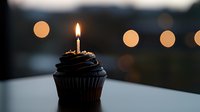 Dark chocolate cupcake with single burning candle against blurred bokeh lights background creates moody celebratory atmosphere for birthday or special occasion.
