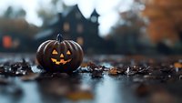 Glowing carved pumpkin with classic jack-o-lantern face sits among fallen autumn leaves at dusk, with spooky silhouette of gothic house in blurred background.